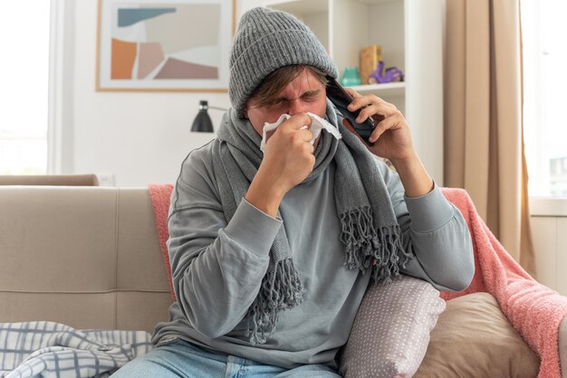 sick young man with scarf around neck wearing winter hat wipes his nose and talking on phone sitting on couch at living room