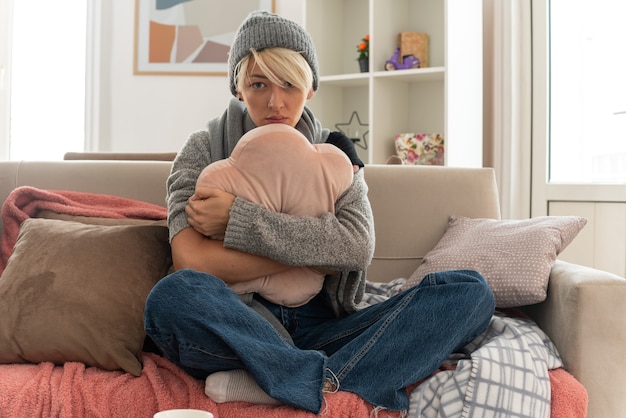 sick young slavic woman with scarf around her neck wearing winter hat hugging pillow sitting on couch at living room