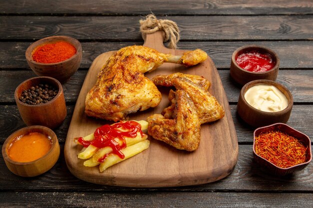 Side close-up view chicken and spices chicken wings and leg french fries and ketchup on the cutting board between bowls of colorful spices and sauces on the dark table