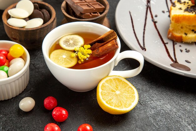 Side close-up view a cup of tea a cup of tea with lemon and cinnamon sticks plate of cake and bowls of chocolate and candies on the dark table