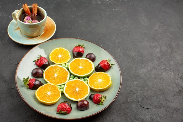 Free photo side close-up view cup of tea and fruits a cup of tea with cinnamon sticks next to the plate of fruits on the black table