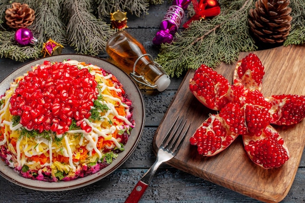 Side close-up view dish and spices dish with pomegranate potatoes next to the bottle of oil pilled pomegranate fork on the cutting board and tree branches with cones and Christmas tree toys