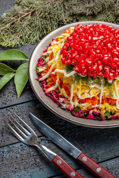 Free photo side close-up view dish with pomegranates dish with pomegranates and fork with knife next to the spruce branches on the dark table