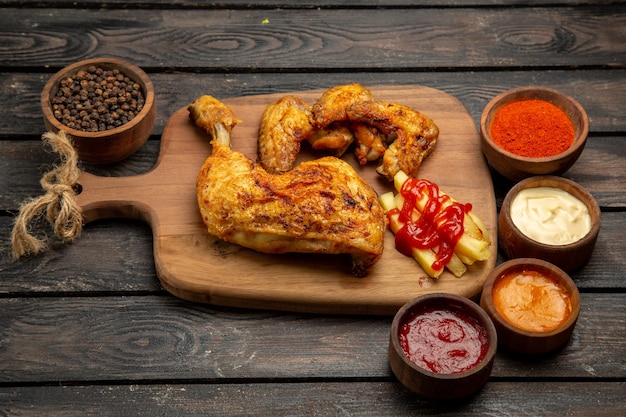 Side close-up view fastfood chicken and french fries on the kitchen board next to the ketchup black pepper sauces and spices on the table