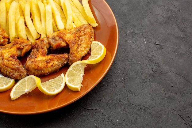 Side close-up view fastfood orange plate of an appetizing chicken wings french fries and lemon on the dark background