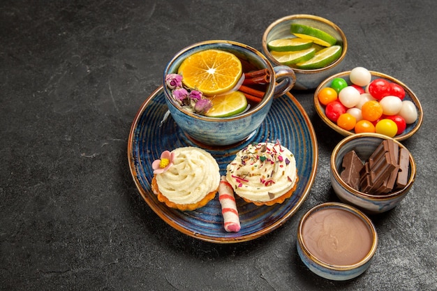 Free photo side close-up view herbal tea a cup of tea with lemon and cupcakes with cream next to the bowls of chocolate slices of lime chocolate cream and colorful sweets on the table