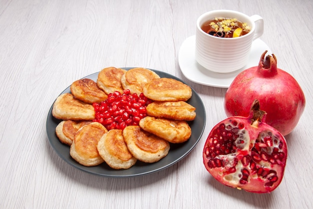 Side close-up view pancakes black plate of pancakes and seeds of pomegranate a cup of herbal tea and pomegranate on the table