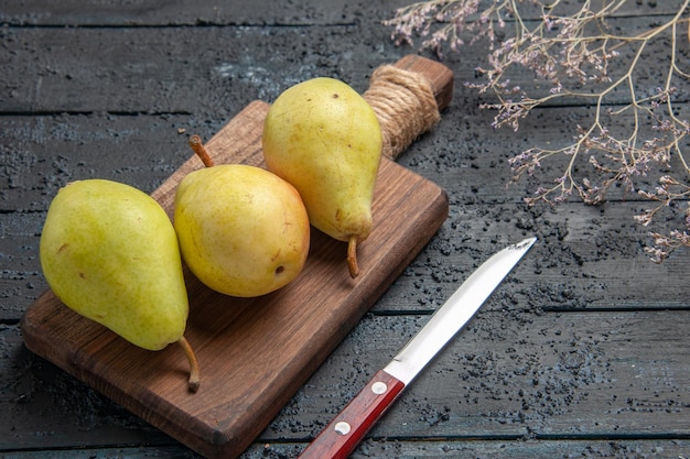 Side close-up view pears on board three green-yellow-red pears on cutting board in the center of dark table next to knife and tree branches