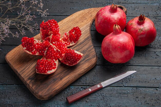 Side close-up view pomegranates knife board ripe pilled pomegranate on cutting board next to three red pomegranates knife and tree branches on dark background