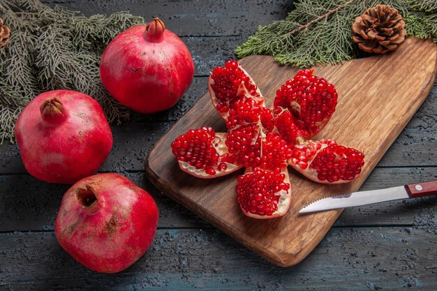 Side close-up view red pomegranates pilled pomegranate on kitchen board next to branches with cones knife and three pomegranates on grey table