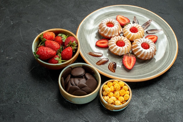 Side close-up view sweets and cookies appetizing cookies with strawberry and bowls of hizelnuts chocolate and strawberry on black table