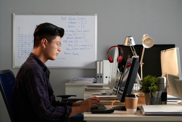 Side shot of young Asian man in glasses working on computer in office