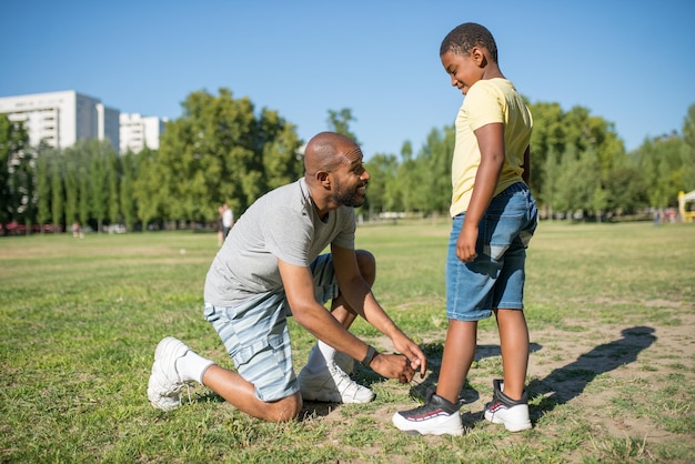 Side view of African dad and his son standing on field. Smiling man standing on his one knee on grass tying shoelaces on kid sneakers both looking at each other. Parents care and sport activity concep