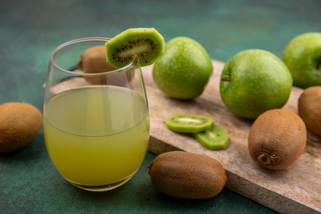 Free photo side view apple juice in a glass with green apples and kiwi on a board  on a green background