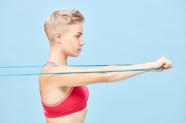 Free photo side view of athletic confident young woman in red top exercising at blue wall using resistance band, pulling it to train arm muscles. strength, energy, determination and motivation concept