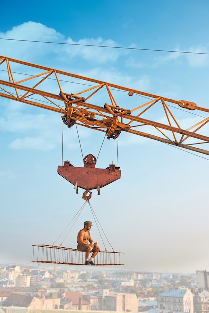 Free Photo side view of athletic man in hat sitting and resting on construction on high and eating. large building crane holding construction with male over city in air. cityscape and blue sky on background.