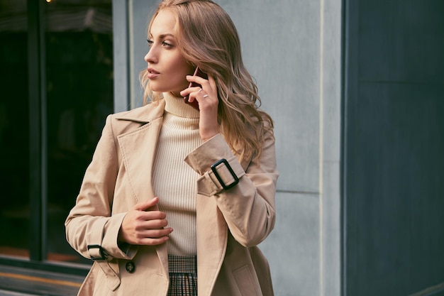 Side view of attractive casual blond girl in trench coat talking on cellphone dreamily looking aside outdoor