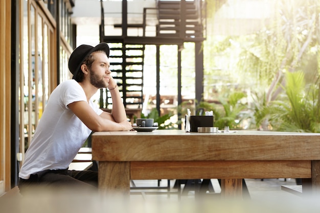 Free Photo side view of attractive young hipster in hat sitting alone at pavement cafeteria, resting his elbow on massive wooden table