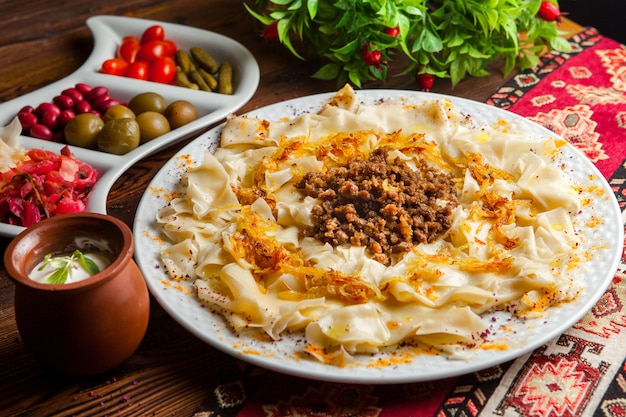 Free photo side view azerbaijani guru khingal caucasian pasta with fried chopped meat and onion with sour cream sauce and pickles on a tablecloth on a dark wooden table horizontal