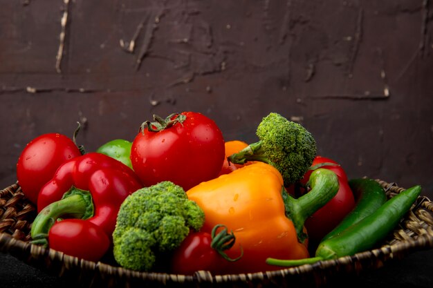 Side view of basket plate full of vegetables as peppers broccoli and tomatoes on black surface and maroon surface