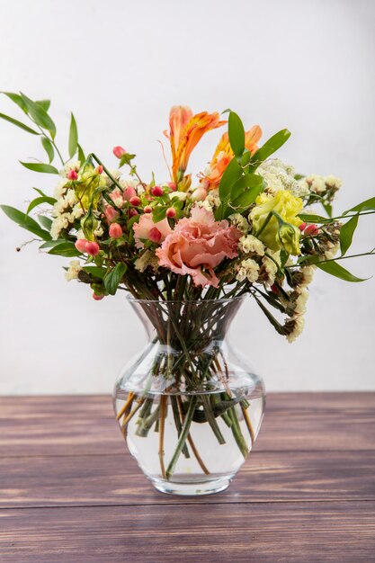 Side view of beautiful different colorful flowers with leaves on a glass vase on a wooden table on white surface