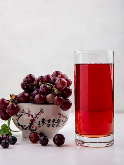 Side view of black grape juice in glass and bowl of red grapes on white background