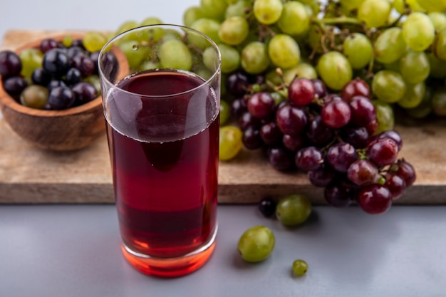 Free photo side view of black grape juice in glass and grapes in bowl and on cutting board on gray background