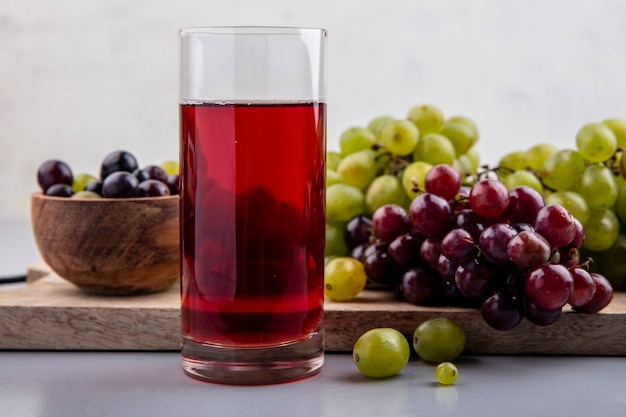 Side view of black grape juice in glass and grapes in bowl and on cutting board on gray surface and white background