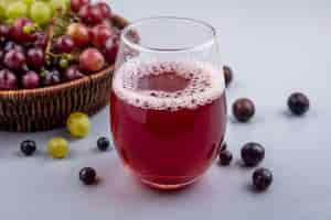 Free photo side view of black grape juice in glass with grapes in basket and on gray background