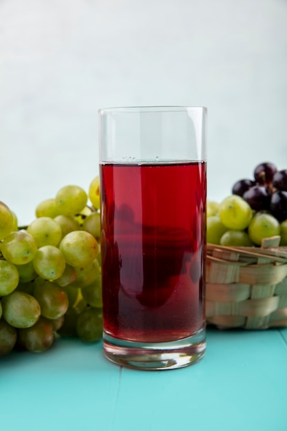 Side view of black grape juice in glass with grapes on blue surface and white background