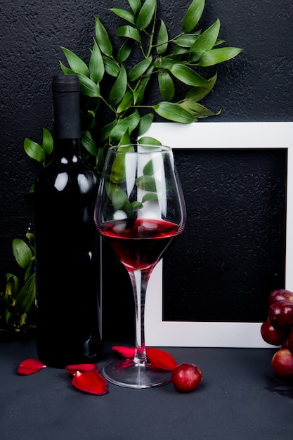 Side view of bottle and glass of red wine with frame and leaves with flower petals on black with copy space