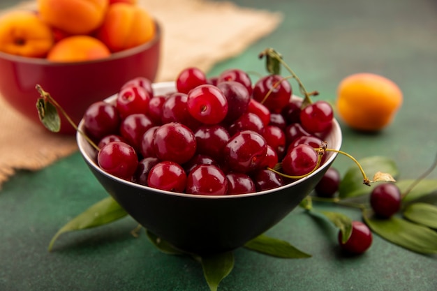 Side view of bowl of cherries with bowl of apricots on sackcloth and leaves on green background