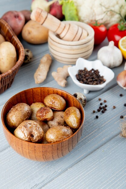 Side view of bowl full of baked potato with vegetables garlic crusher black pepper on wooden background