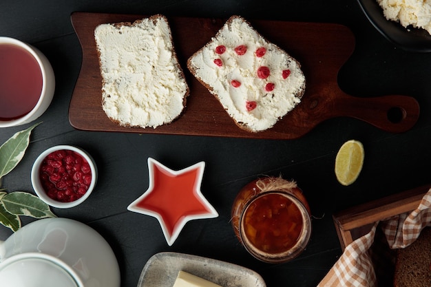 Side view of bread slices smeared with cottage cheese with red currants on it on cutting board with jam butter lime slice red currant on black background