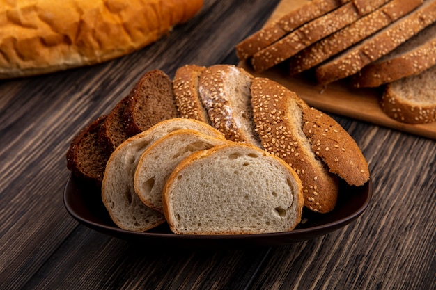 Side view of breads as sliced seeded brown cob rye and white ones in bowl and on cutting board on wooden background