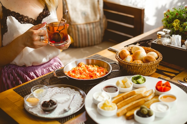 Side view breakfast set azerbaijani style eggs with tomatoes toast jam honey fresh tomato cucumber cheese french bread rolls and black tea in a cup