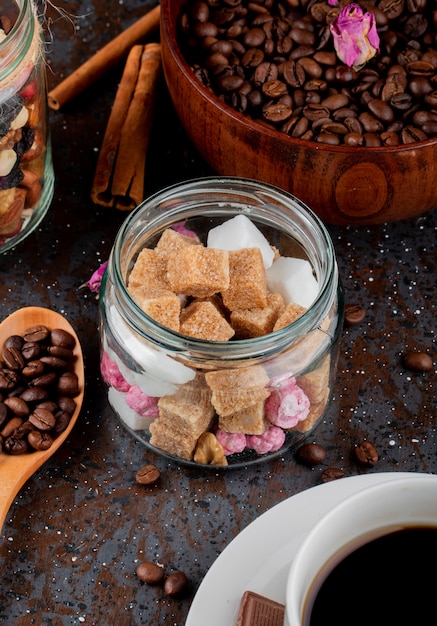 Side view of brown sugar cubes in a glass jar and coffee beans in a bowl on black background