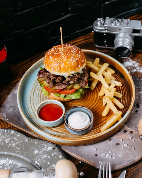 Side view of burger with beef meat pickles and tomatoes served with french fries and sauces on black