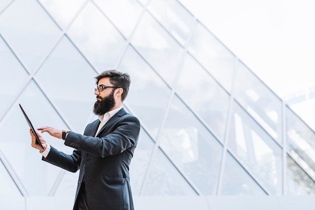 Free photo side view of a businessman with long beard using digital tablet standing against glass building