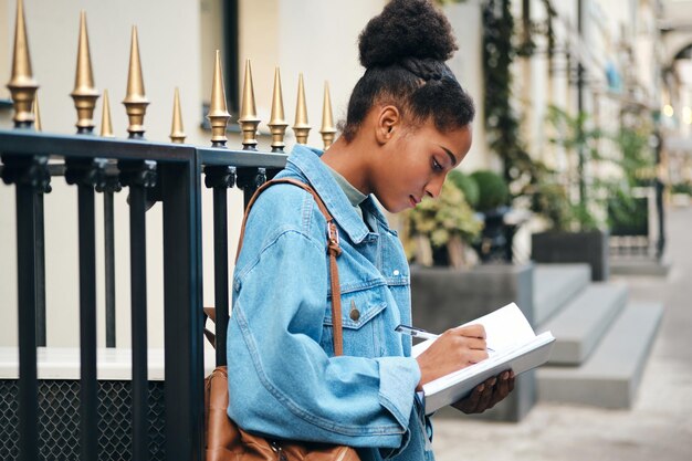 Side view of casual African American student girl in denim jacket with backpack writing notes on city street