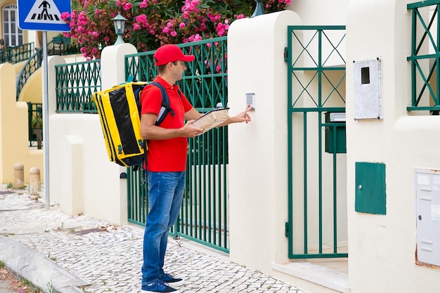 Free photo side view of caucasian courier ringing in doorbell. pensive postman wearing red uniform, carrying yellow thermal bag, holding package and standing outdoors. delivery service and post concept