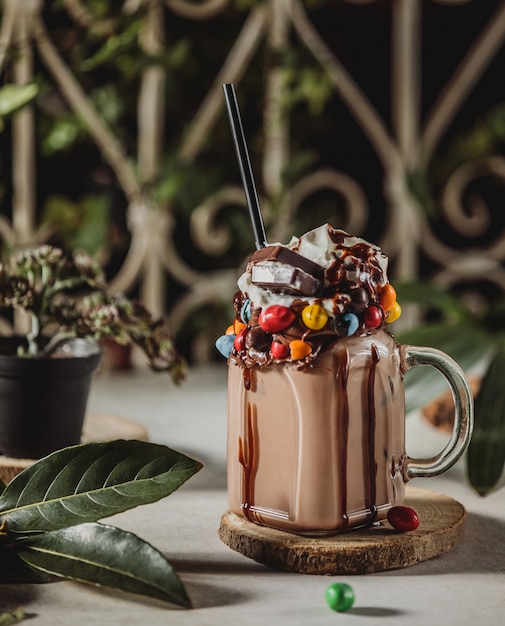 Free photo side view of chocolate milk shake with whipped cream decorated with candies in a glass jar with handle on a wooden stand