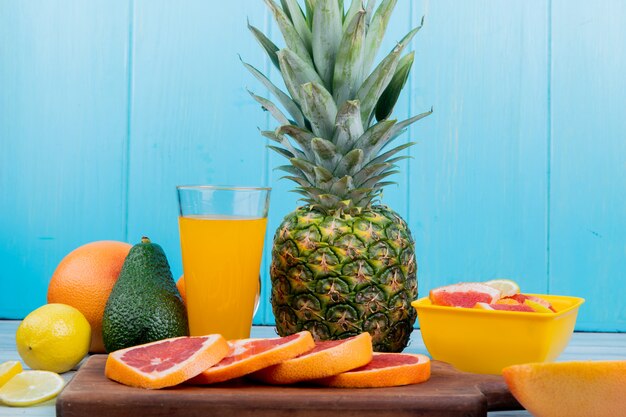 Side view of citrus fruits as lemon avocado pineapple with orange juice and sliced grapefruit on cutting board on wooden surface and blue background