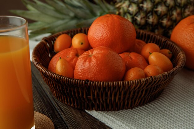 Side view of citrus fruits as tangerines and kumquats in basket orange pineapple on cloth with orange juice on wooden background