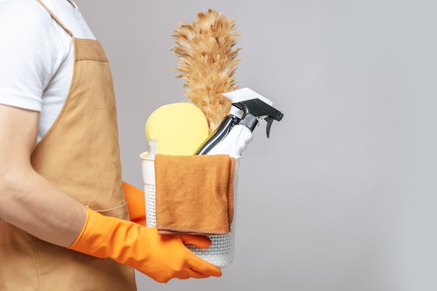 Side view, Close up hand of Young man in apron and rubber gloves holding a basket of cleaning equipment, the feather duster, spray bottle, sponge and cloth for wiping in basket