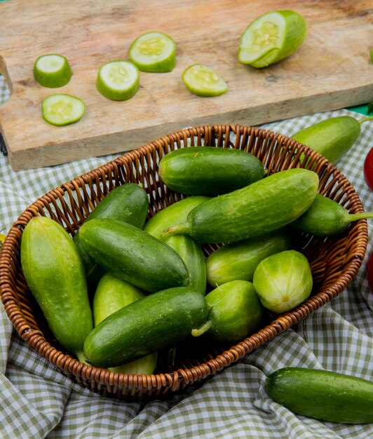 Side view of cucumbers in basket with sliced cucumber on cutting board on plaid cloth