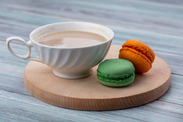 Side view of cup of cappuccino with colored macarons on a wooden stand on a gray surface