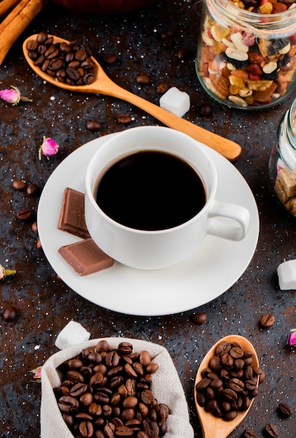 Side view of a cup of coffee with chocolate and a wooden spoon with coffee beans on black background