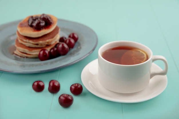 Side view of cup of tea on saucer and pancakes with cherries in plate and on blue background