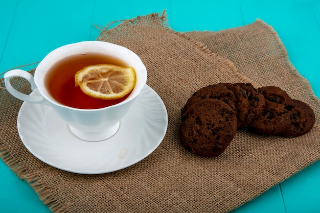 Side view of cup of tea on saucer with slice of lemon and cookies on sackcloth and blue background